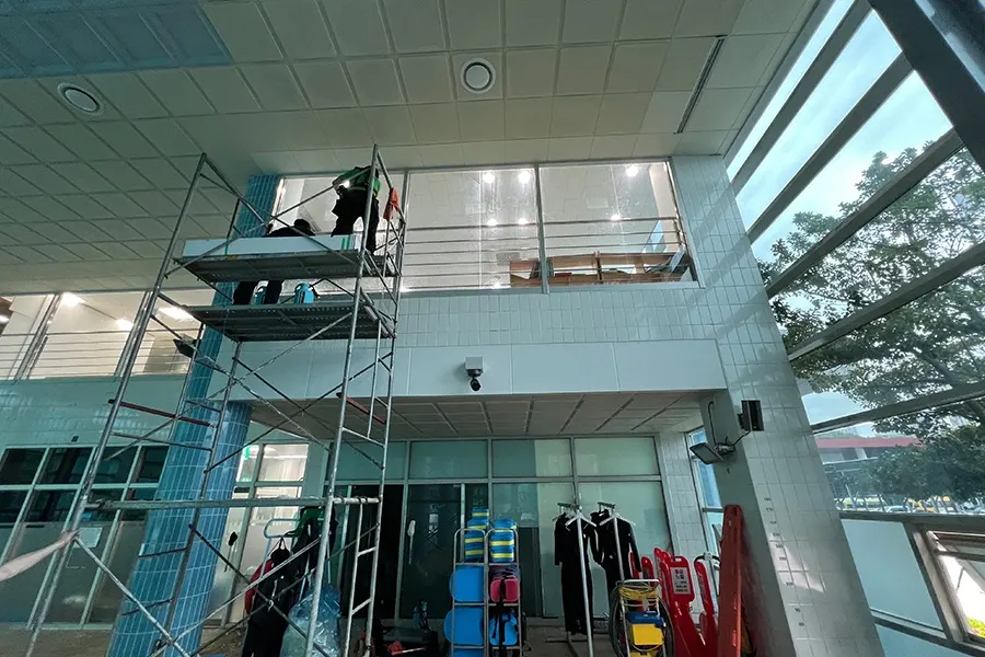 2층 높이 창가에 세운 작업대에 올라 작업 중인 기사들, 오른쪽 유리 너머로 야외 나무가 보이는 모습